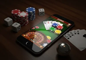 Smartphone showing an online roulette and card game interface surrounded by poker chips, dice, and playing cards on a wooden table, representing online casino gaming.